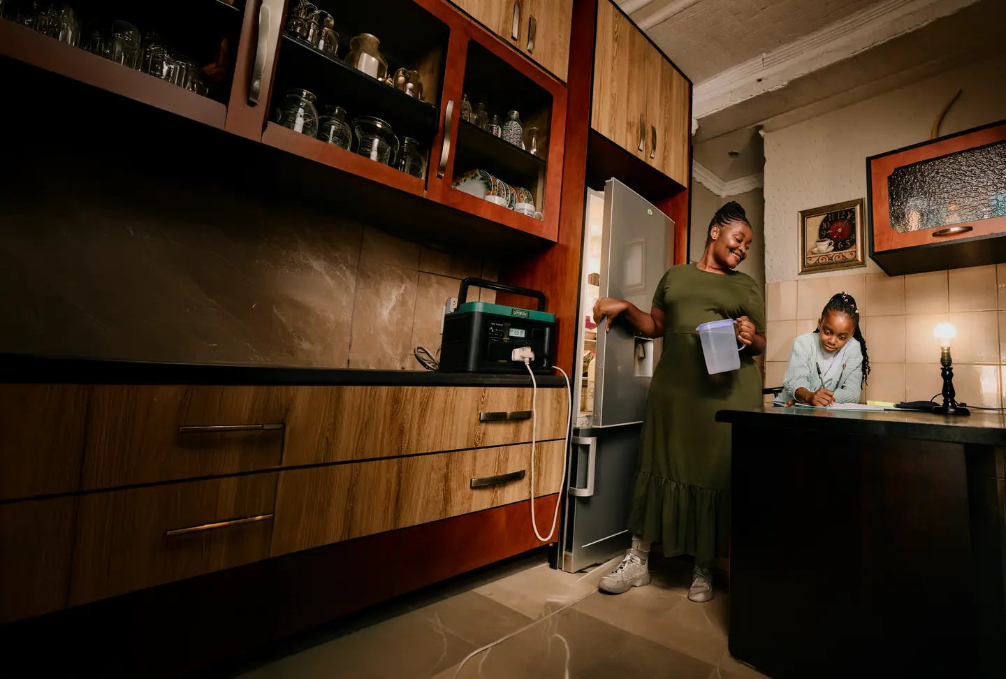 A woman at her fridge and a girl doing homework, both powered by a bPOWERd battery during load shedding